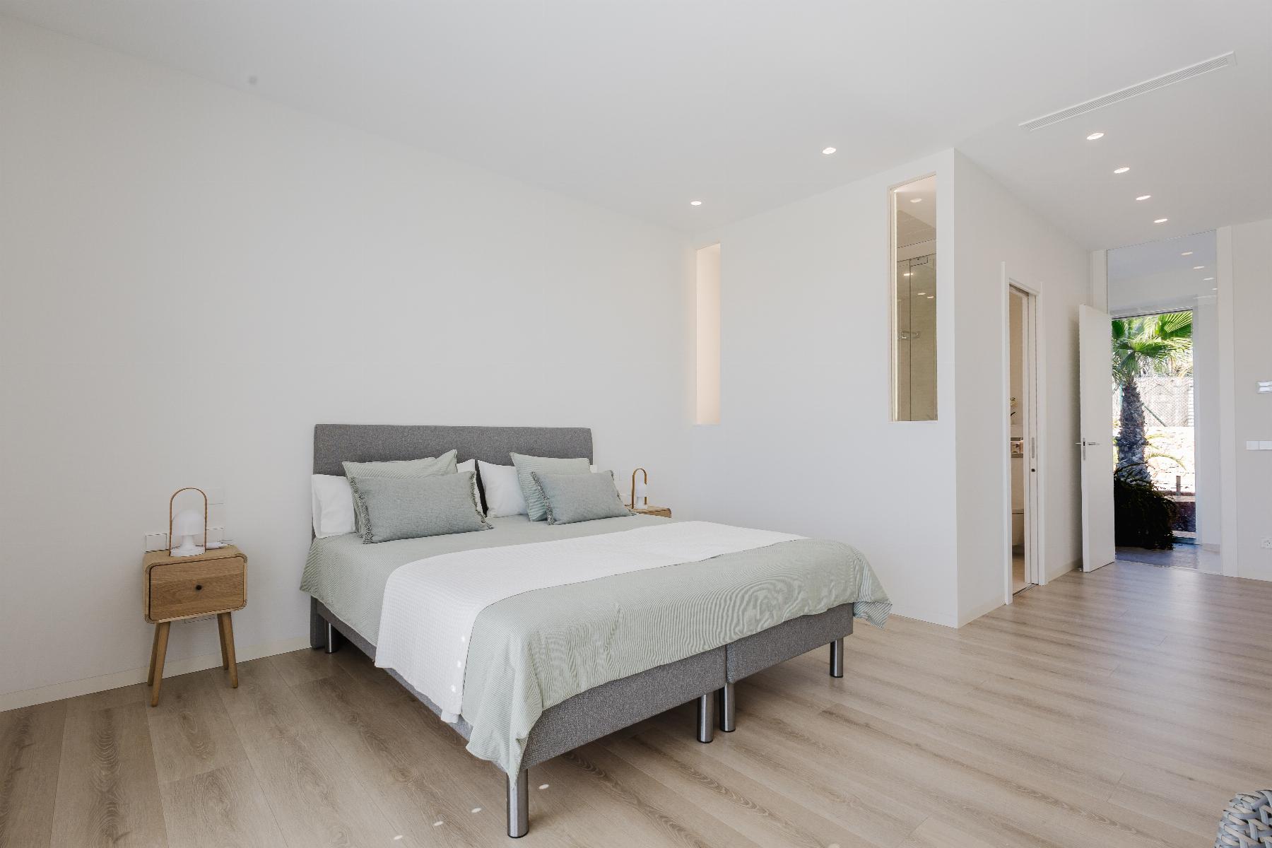 Minimalist bedroom with a gray upholstered bed, pale green bedding, light wood floor, and open doorway to outdoors.