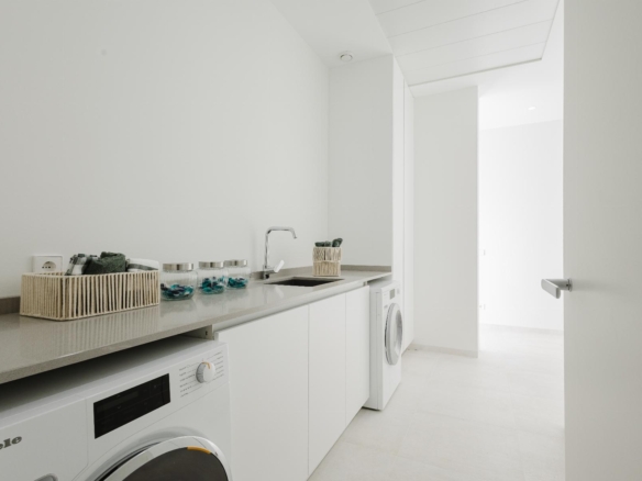 Bright white laundry room with a long counter, sink, and a row of washing machines underneath. A wicker basket and glass jars sit on the counter with towels inside a smaller basket in the background.