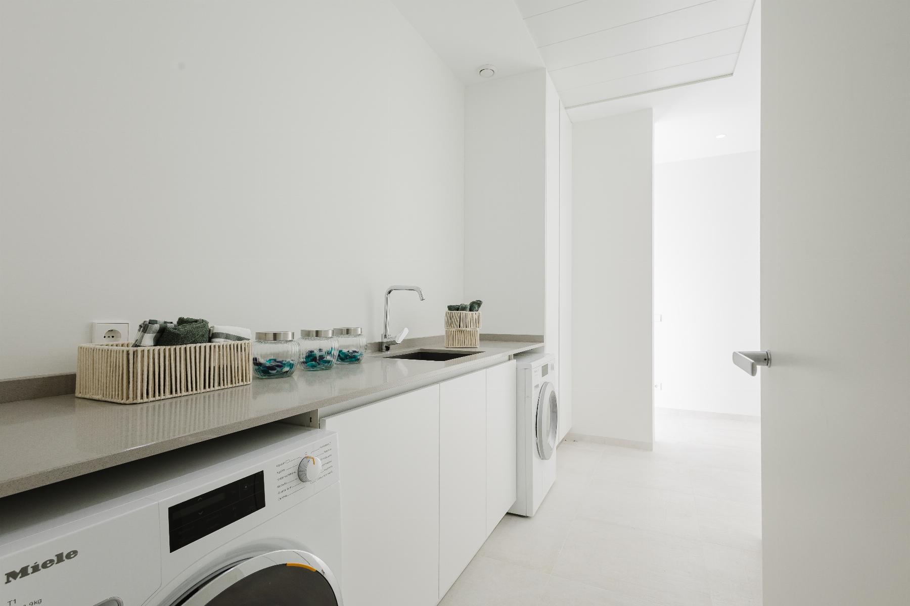 Bright white laundry room with a long counter, sink, and a row of washing machines underneath. A wicker basket and glass jars sit on the counter with towels inside a smaller basket in the background.
