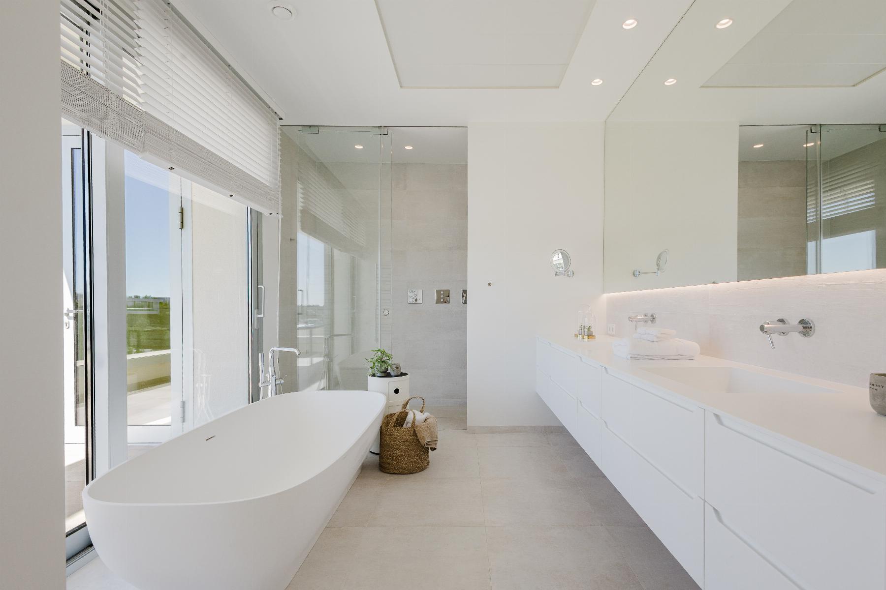 Bright minimalist bathroom with a freestanding oval tub, glass shower, and long white vanity beneath bright windows.