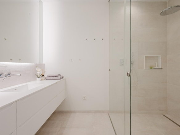 Minimalist white bathroom with a long counter and sink on the left, wall-mounted faucet, and folded towels; glass shower enclosure on the right.