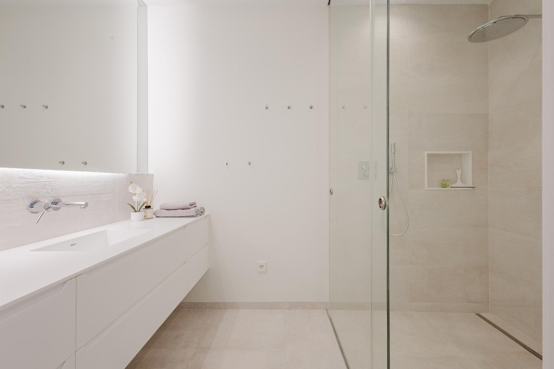 Minimalist white bathroom with a long counter and sink on the left, wall-mounted faucet, and folded towels; glass shower enclosure on the right.