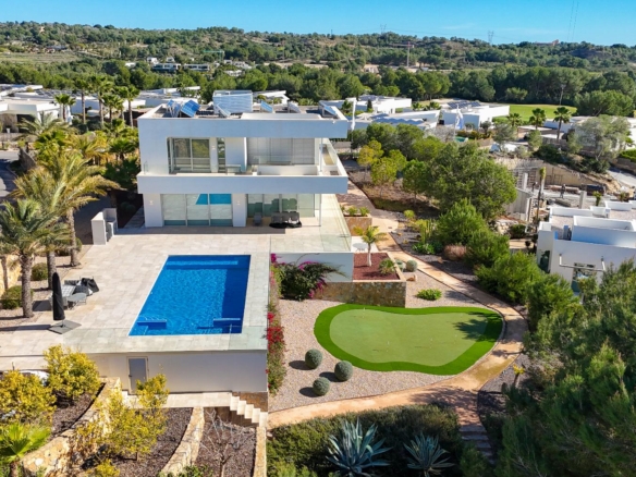 Aerial view of a modern white villa with a rectangular blue pool, lounge area, and a manicured putting green in a landscaped yard.