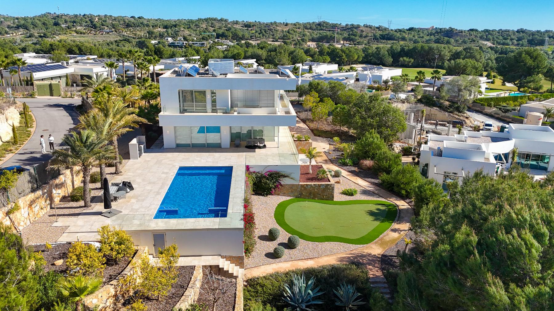 Aerial view of a modern white villa with a rectangular blue pool, lounge area, and a manicured putting green in a landscaped yard.