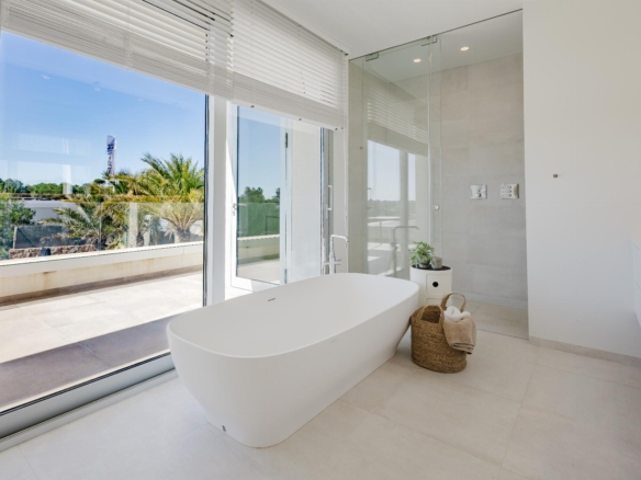 Bright modern bathroom with a freestanding white tub, glass shower enclosure, and large windows overlooking a patio with palm trees on a sunny day.