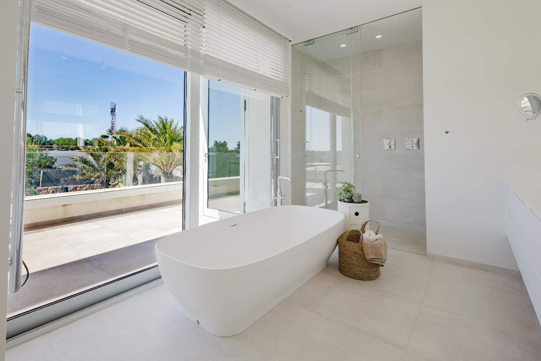 Bright modern bathroom with a freestanding white tub, glass shower enclosure, and large windows overlooking a patio with palm trees on a sunny day.