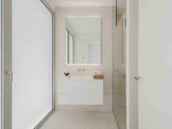 Minimalist bathroom with a floating white vanity, backlit mirror, frosted glass door on the left, and a glass shower enclosure on the right; towels and a small plant on the counter.