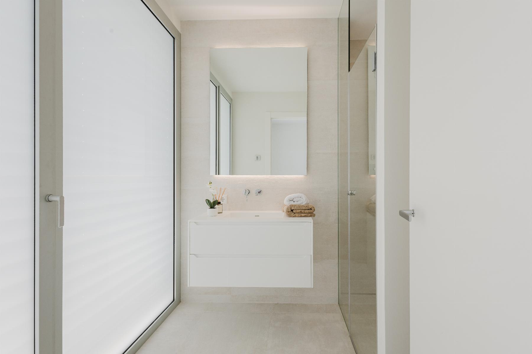 Minimalist bathroom with a floating white vanity, backlit mirror, frosted glass door on the left, and a glass shower enclosure on the right; towels and a small plant on the counter.