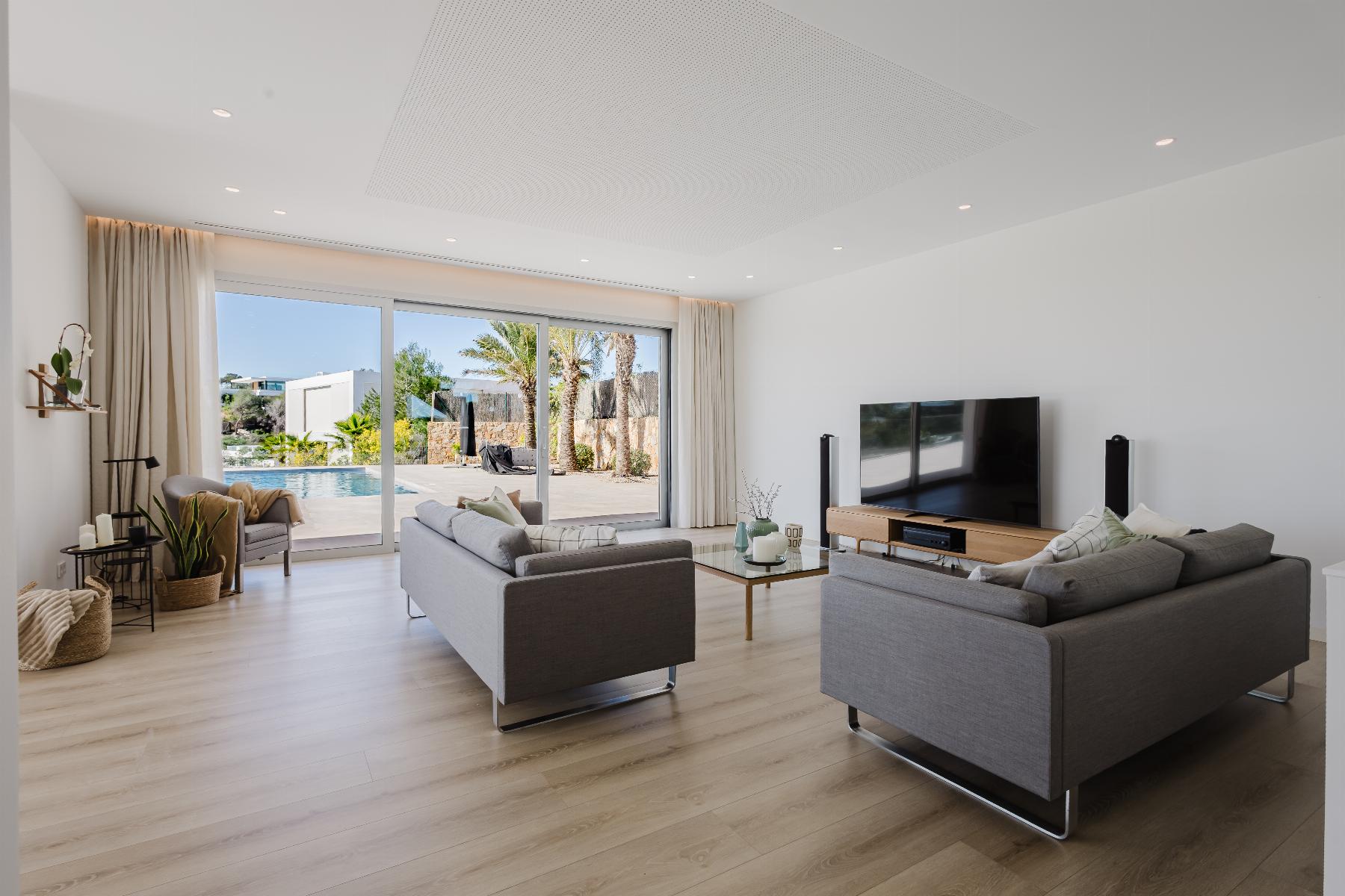 Bright contemporary living room with gray sofas, a large TV, and sliding glass doors opening to a sunny backyard and pool area.