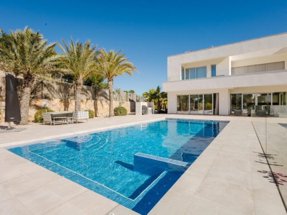Modern white two-story villa with glass balconies, a blue swimming pool in the foreground, and palm trees along a stone wall under a clear blue sky.