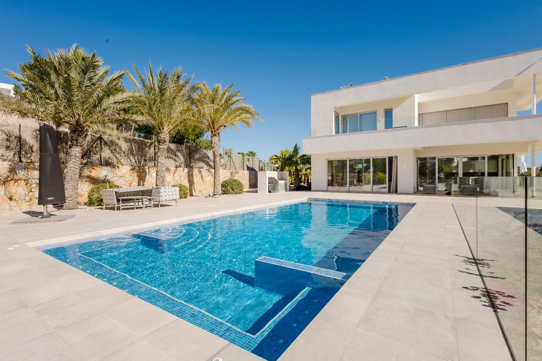 Modern white two-story villa with glass balconies, a blue swimming pool in the foreground, and palm trees along a stone wall under a clear blue sky.