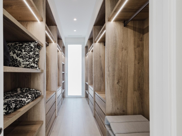 Walk-in closet with light wood shelves and drawers on both sides, LED lighting, and a cushioned bench at the end.