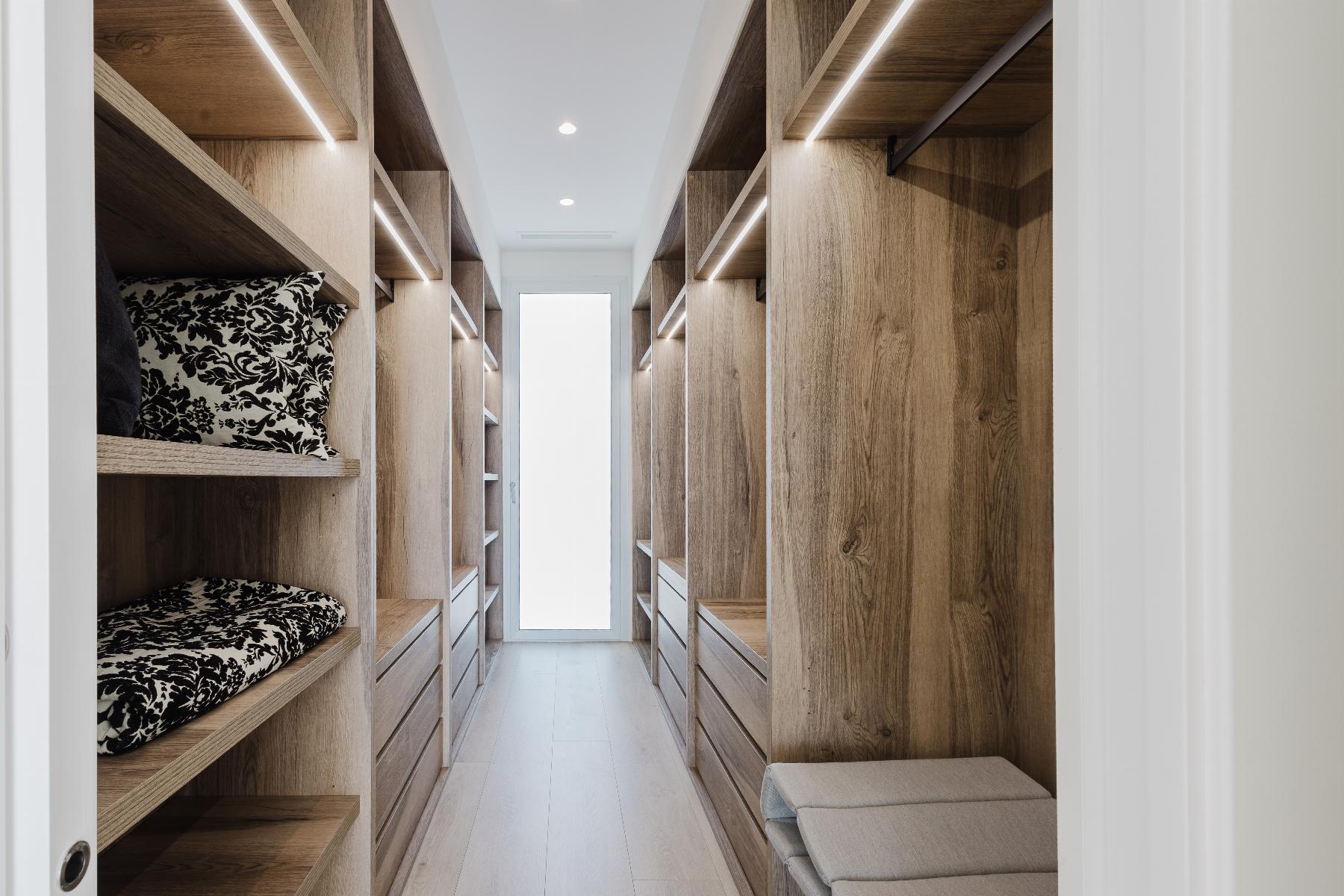 Walk-in closet with light wood shelves and drawers on both sides, LED lighting, and a cushioned bench at the end.