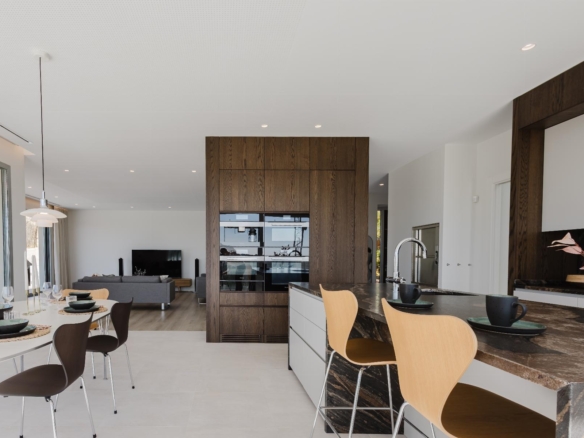 Open-concept kitchen and dining area with a dark marble island, wooden cabinets, and a glass-front pantry; dining table set for a meal and a living room visible in the background.