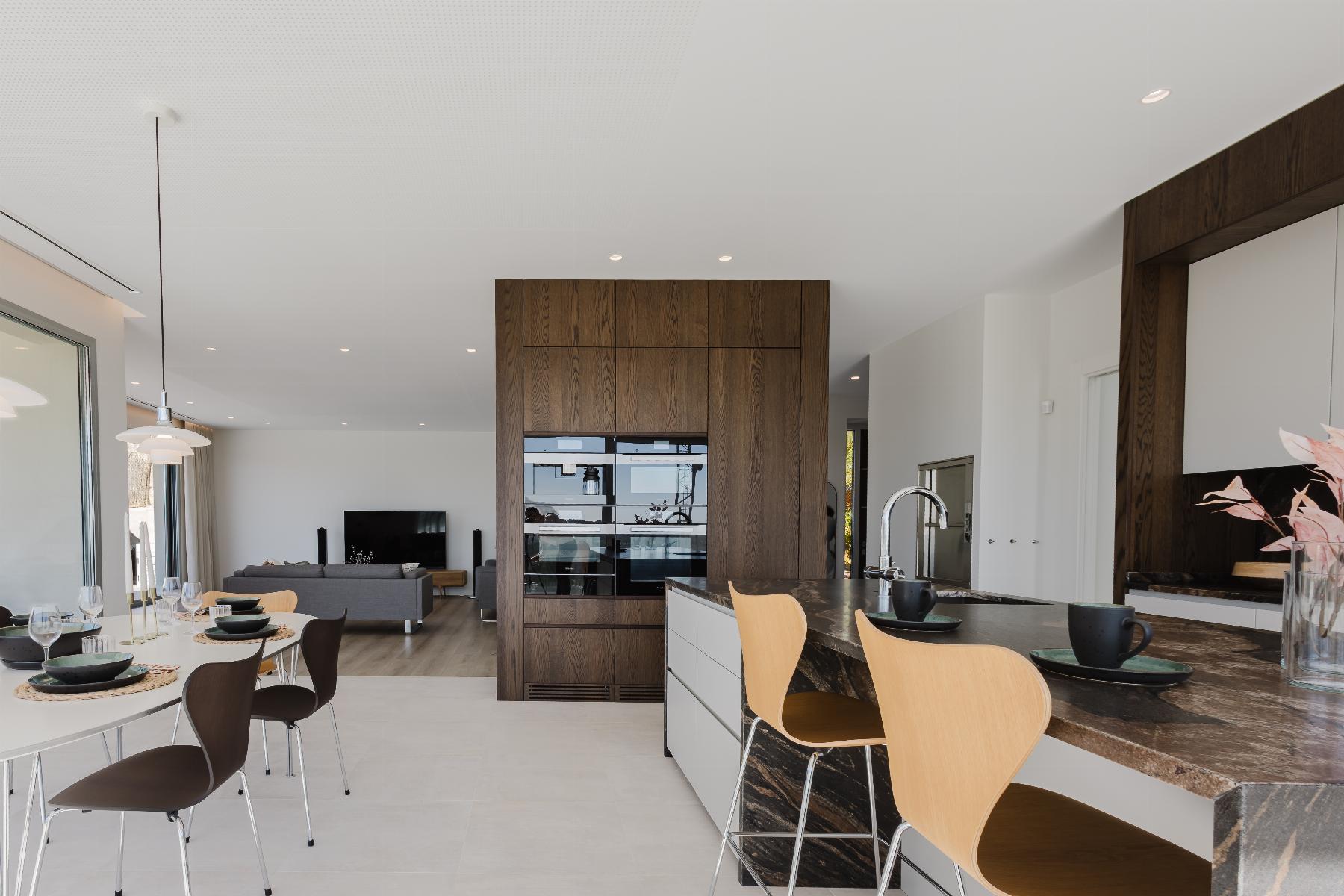 Open-concept kitchen and dining area with a dark marble island, wooden cabinets, and a glass-front pantry; dining table set for a meal and a living room visible in the background.