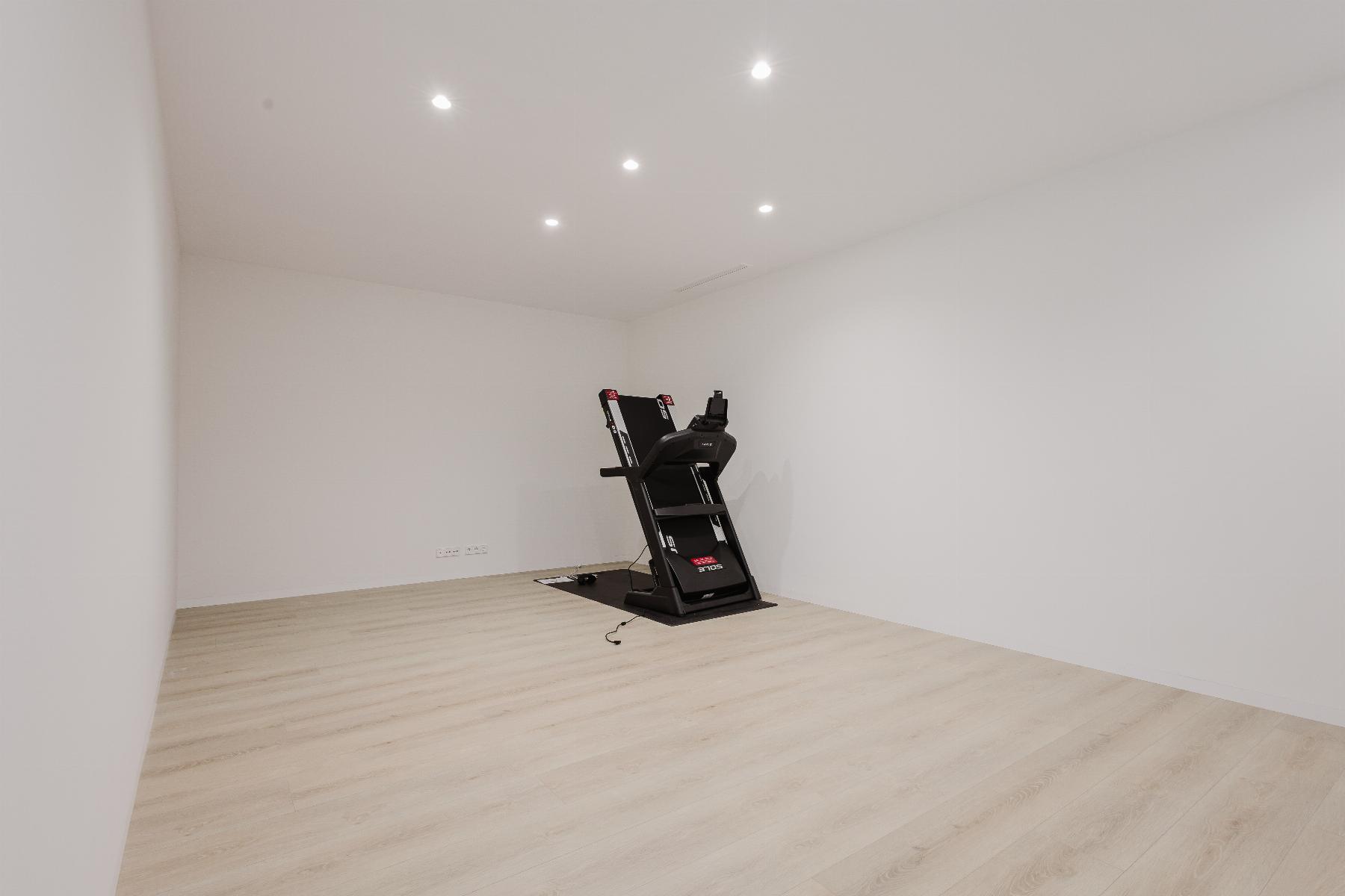 Minimal home gym: white-walled room with light wood floor and a black treadmill on a mat against the back wall.