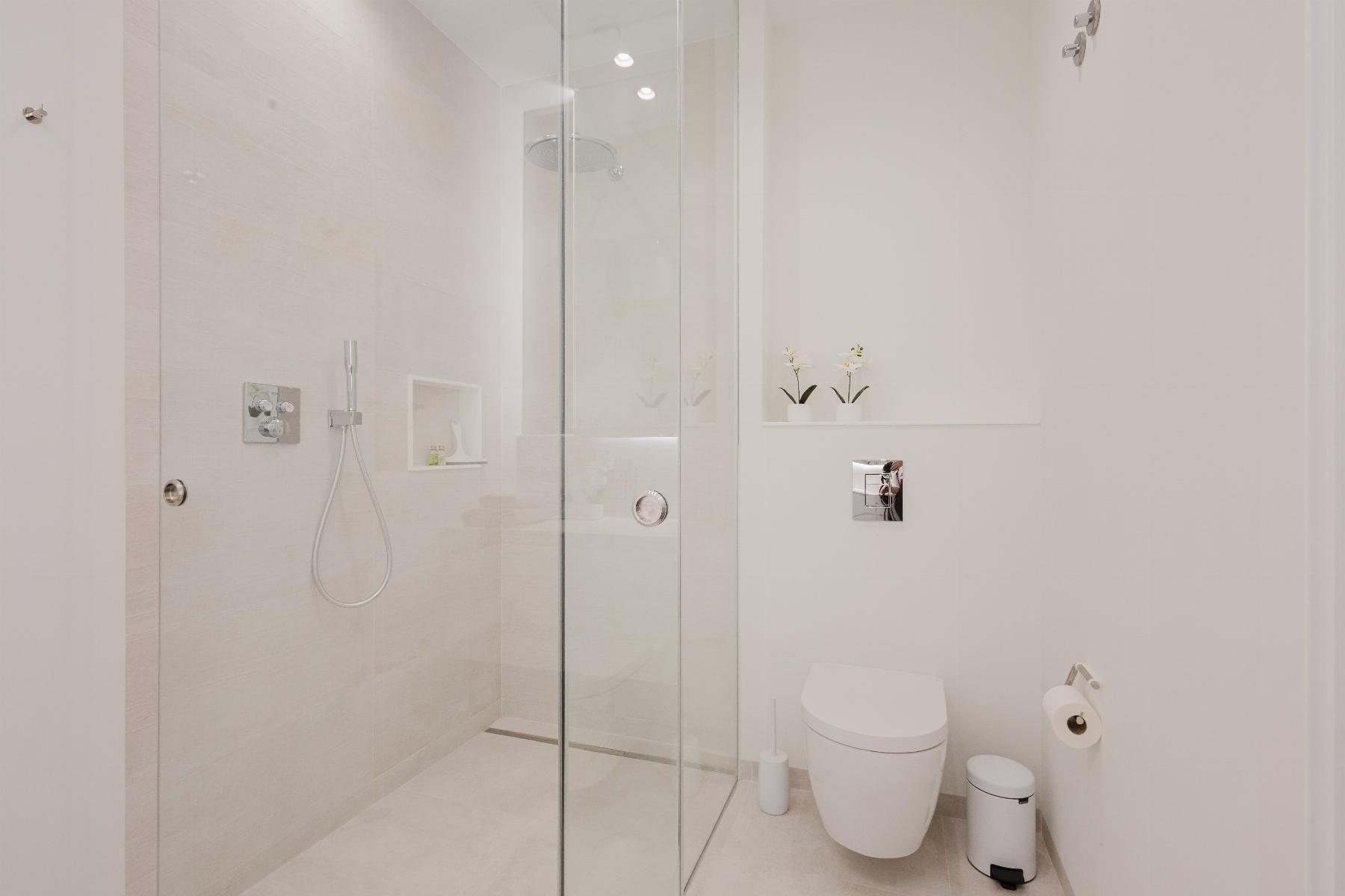 Modern white bathroom with a glass shower enclosure, wall-mounted shower and toilet, plus small potted orchids on a shelf.