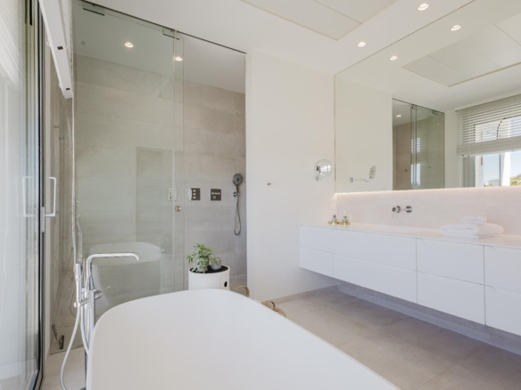 Bright, modern bathroom with a glass shower enclosure, freestanding tub in foreground, and white vanity along a light tiled wall.