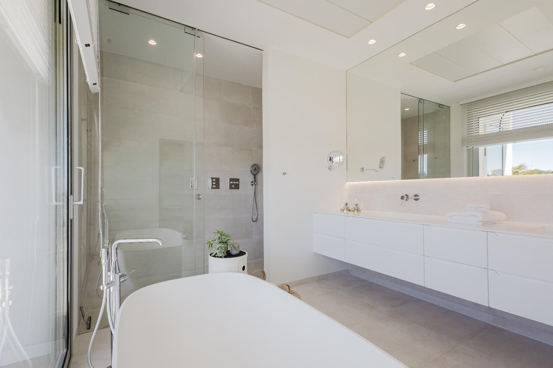 Bright, modern bathroom with a glass shower enclosure, freestanding tub in foreground, and white vanity along a light tiled wall.