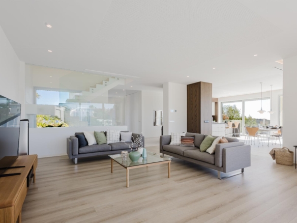 Bright open-plan living room with gray sofas, glass partition, and a sunny kitchen/dining area in the background.