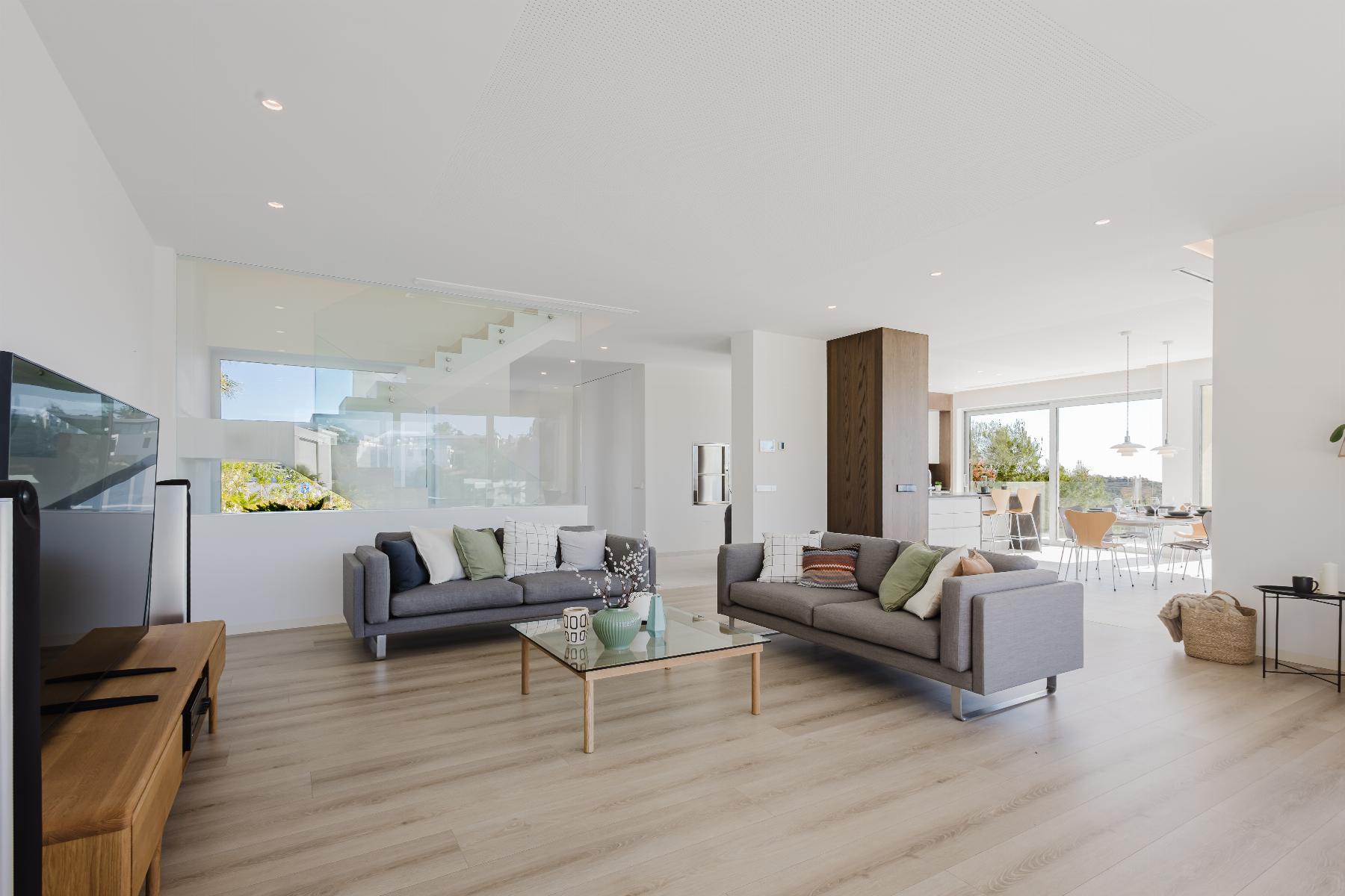 Bright open-plan living room with gray sofas, glass partition, and a sunny kitchen/dining area in the background.