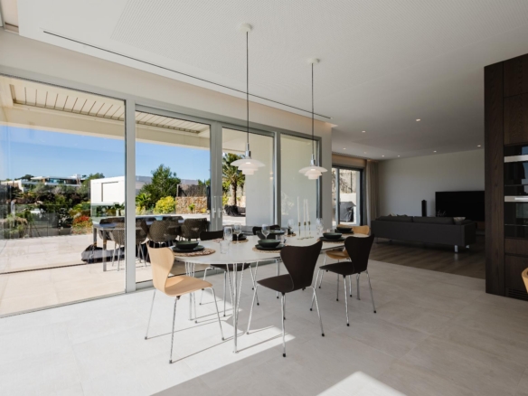 Open-plan dining area with a long white table set with plates and glasses, facing large glass doors onto an outdoor patio. Modern pendant lights hang from the ceiling; adjacent kitchen and living area visible.