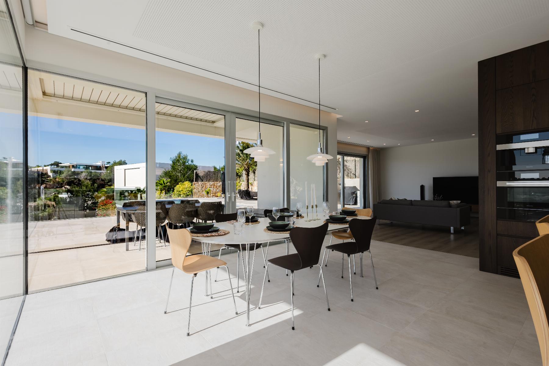 Open-plan dining area with a long white table set with plates and glasses, facing large glass doors onto an outdoor patio. Modern pendant lights hang from the ceiling; adjacent kitchen and living area visible.