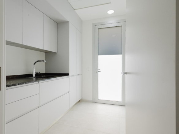 Narrow white kitchen with handle-less cabinets, granite counter, and a curved faucet under a frosted door/window at the far end.