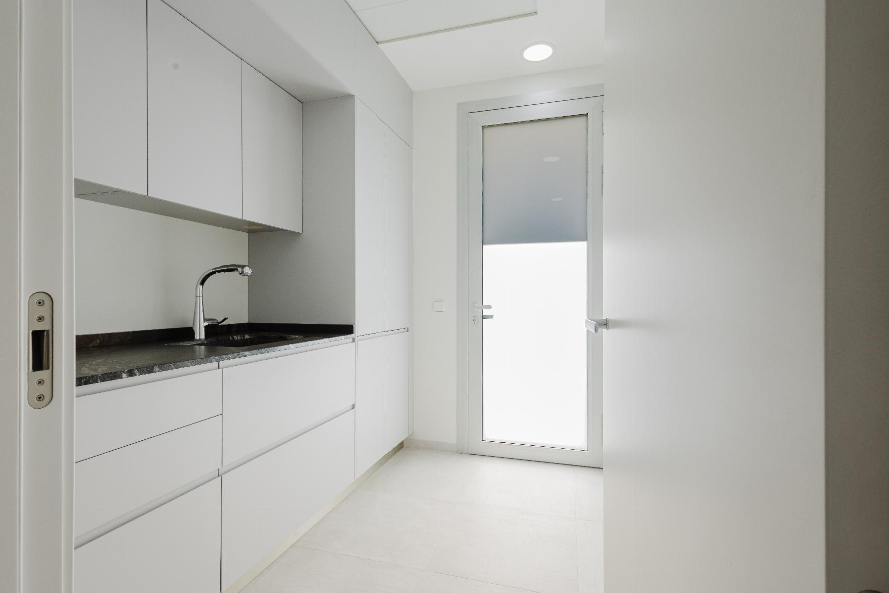 Narrow white kitchen with handle-less cabinets, granite counter, and a curved faucet under a frosted door/window at the far end.