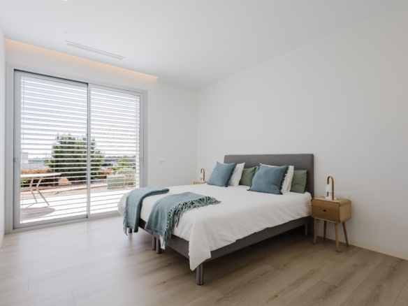 Minimalist bedroom with a gray upholstered bed, white linens, teal pillows, and light wood nightstands; large sliding glass door with blinds opens to a sunny patio.