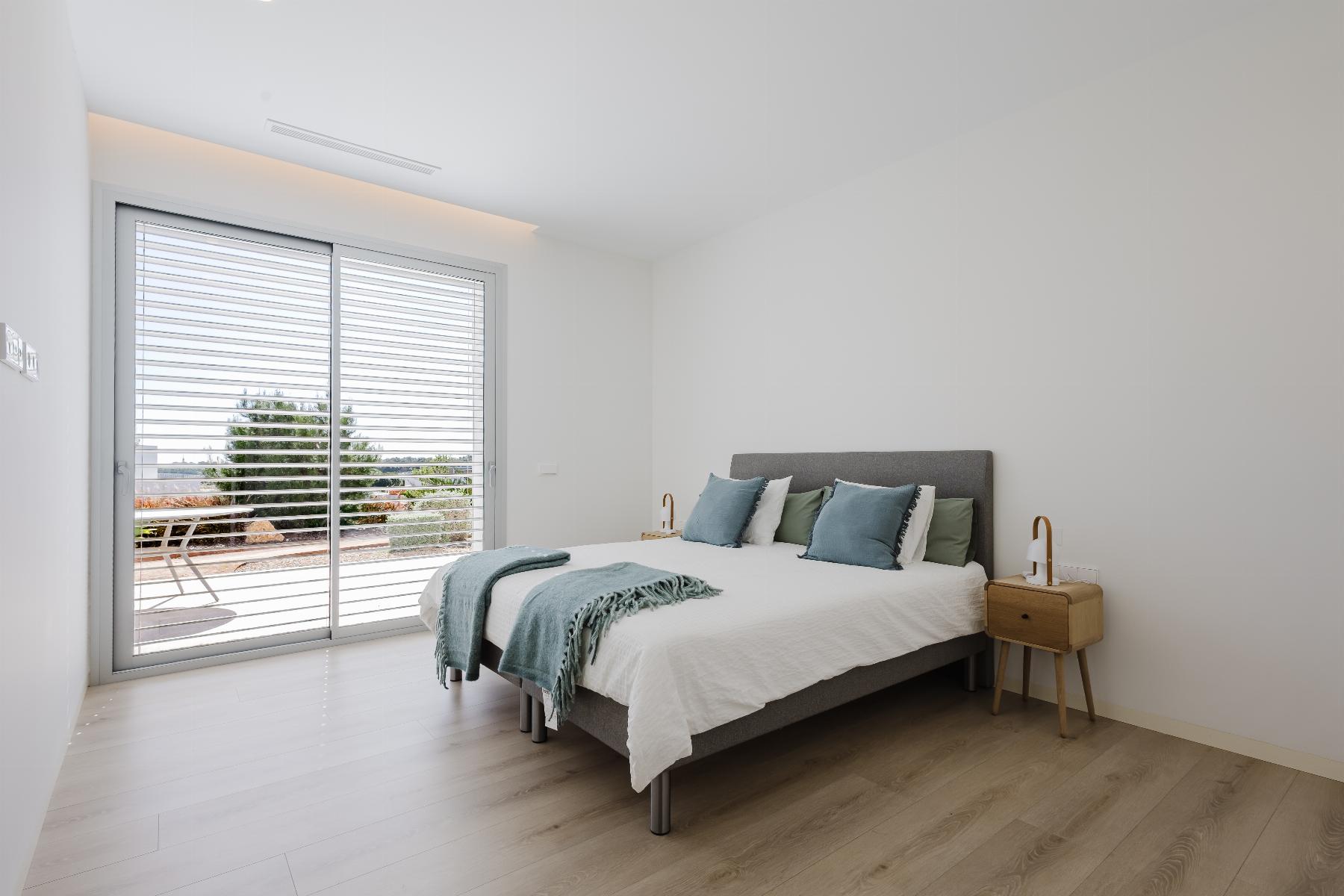 Minimalist bedroom with a gray upholstered bed, white linens, teal pillows, and light wood nightstands; large sliding glass door with blinds opens to a sunny patio.