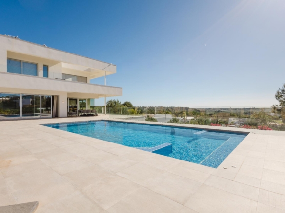 Modern white villa with a rectangular blue pool on a tiled terrace and glass balcony railings under a clear blue sky