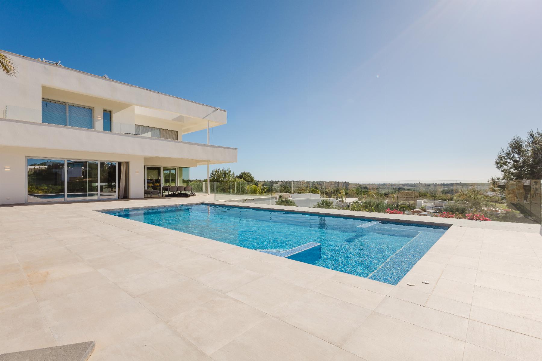 Modern white villa with a rectangular blue pool on a tiled terrace and glass balcony railings under a clear blue sky