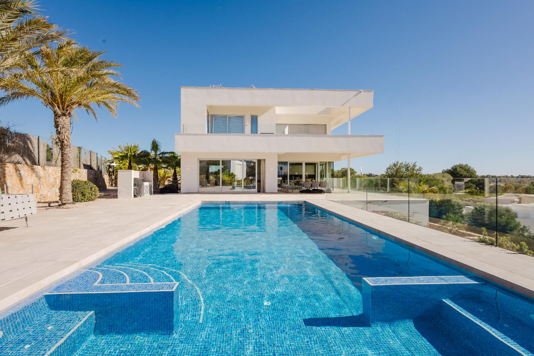 Contemporary white two‑story house with a large rectangular pool in the foreground and palm trees under a clear blue sky.
