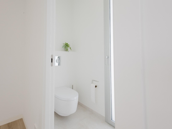 Minimalist white bathroom with a wall-mounted toilet, toilet paper holder, and a small green plant on a shelf in the corner.
