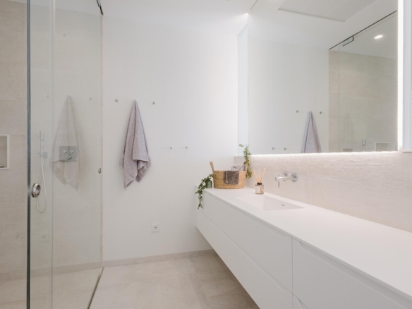 Bright white modern bathroom with a glass shower enclosure, long white vanity, and a large backlit mirror reflecting lavender towels on hooks.