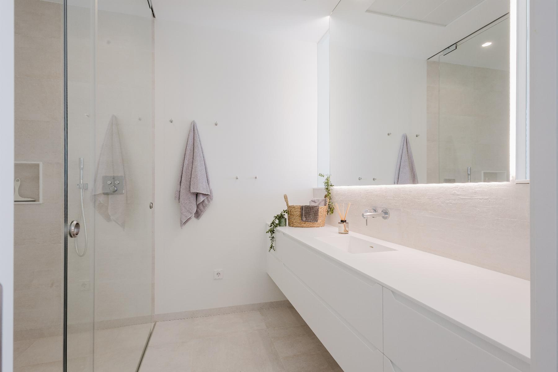 Bright white modern bathroom with a glass shower enclosure, long white vanity, and a large backlit mirror reflecting lavender towels on hooks.