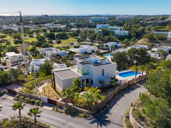Aerial view of a modern hillside residential area with white cubic houses, a construction crane, and a distant golf course under a clear blue sky.