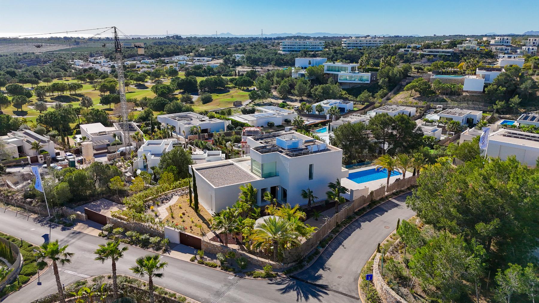 Aerial view of a modern hillside residential area with white cubic houses, a construction crane, and a distant golf course under a clear blue sky.