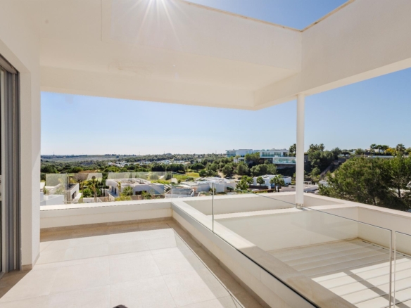 Sunlit white balcony with glass railings offering a panoramic view of a suburban landscape and green trees in the distance.