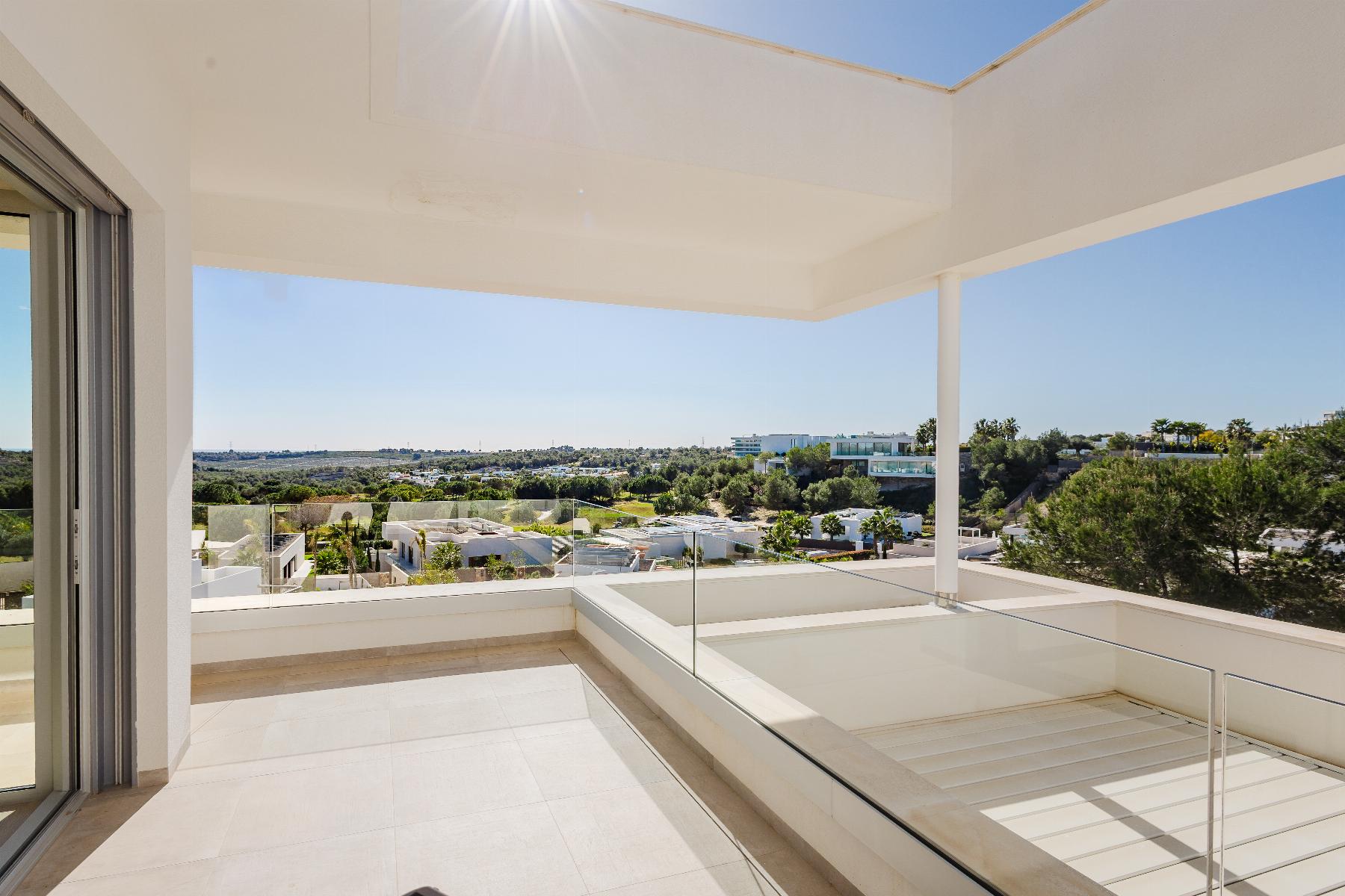 Sunlit white balcony with glass railings offering a panoramic view of a suburban landscape and green trees in the distance.