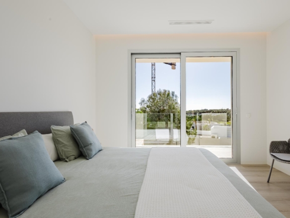 Modern bedroom with a gray upholstered bed, blue-green pillows, and a sliding glass door opening to a balcony with trees and a distant crane.