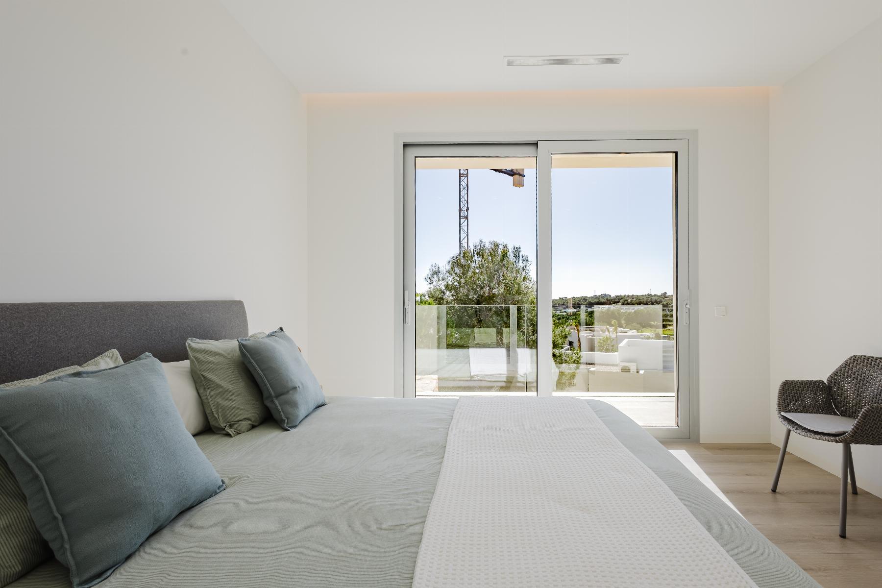 Modern bedroom with a gray upholstered bed, blue-green pillows, and a sliding glass door opening to a balcony with trees and a distant crane.