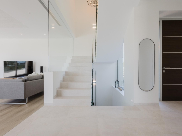 Bright modern living area with a white staircase behind a glass railing, gray sofa, and a wall-mounted TV.