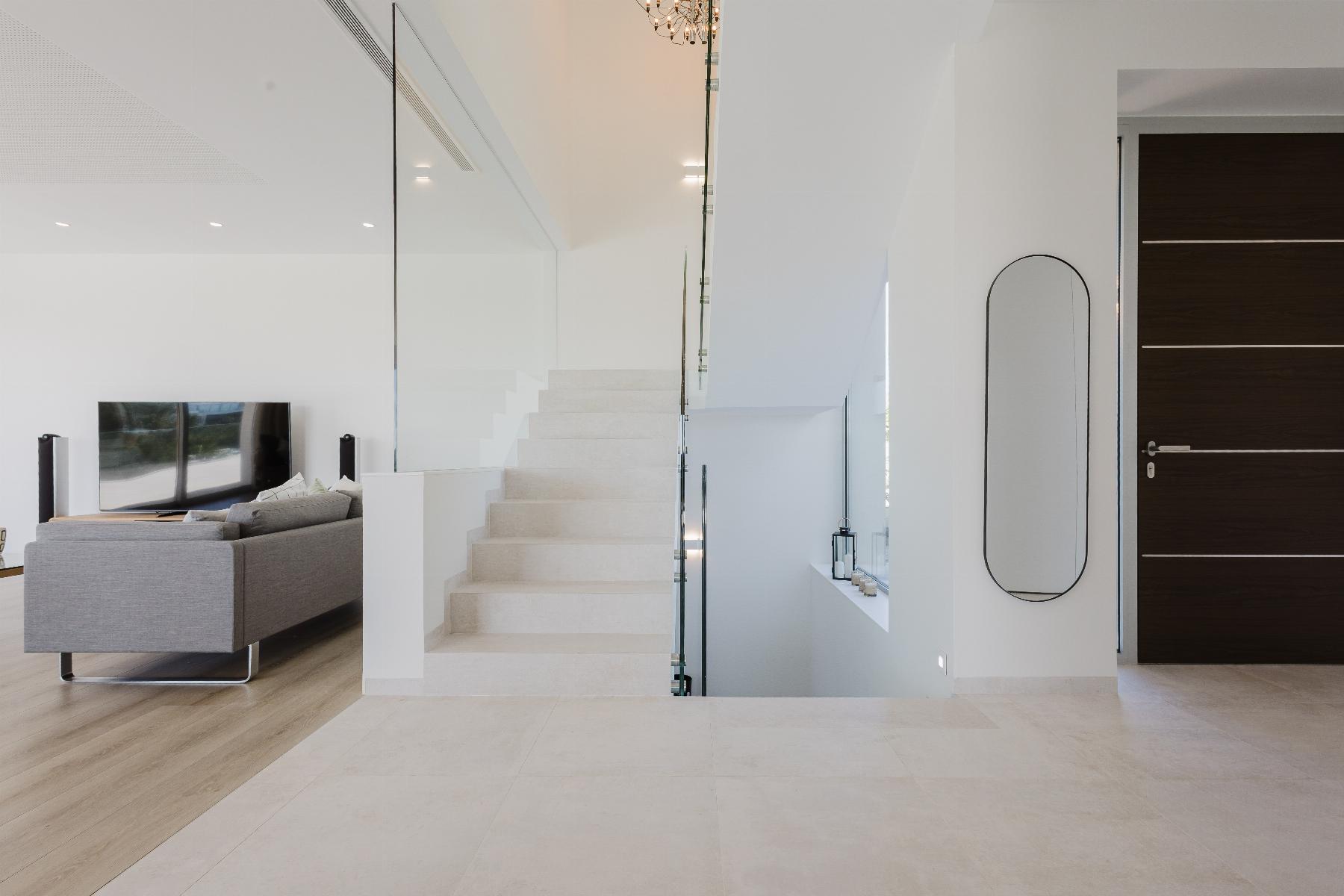 Bright modern living area with a white staircase behind a glass railing, gray sofa, and a wall-mounted TV.