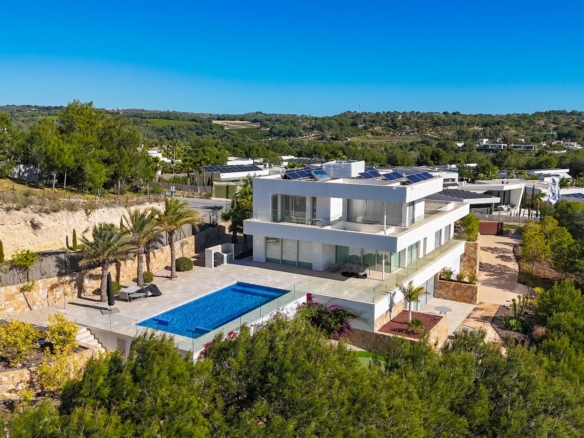 Aerial view of a modern white villa with a rectangular blue pool, glass walls, and a sunlit terrace surrounded by trees.