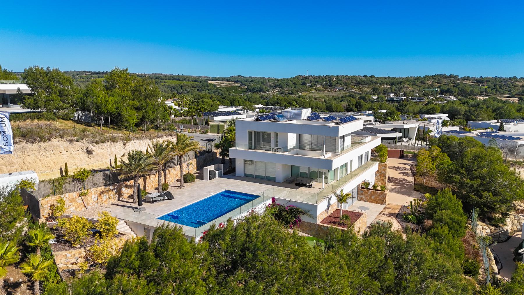 Aerial view of a modern white villa with a rectangular blue pool, glass walls, and a sunlit terrace surrounded by trees.