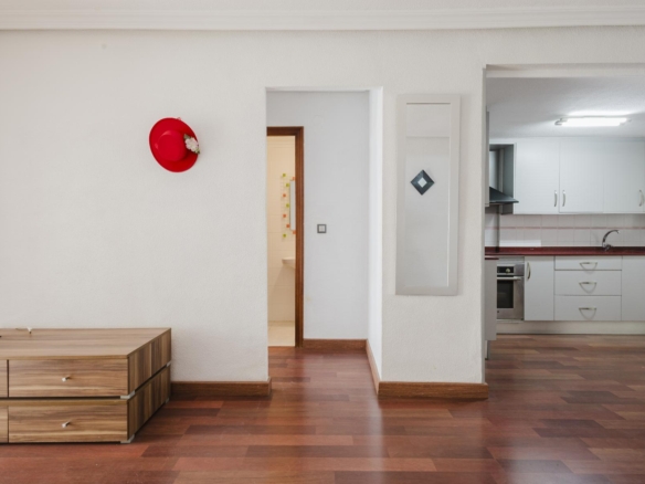 Red hat hanging on a white wall to the left of a doorway; a wooden dresser sits on the left foreground; kitchen on the right.