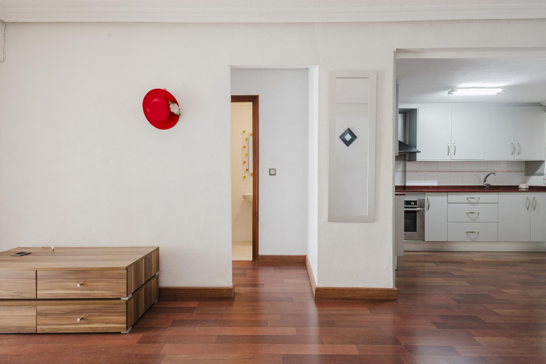 Red hat hanging on a white wall to the left of a doorway; a wooden dresser sits on the left foreground; kitchen on the right.
