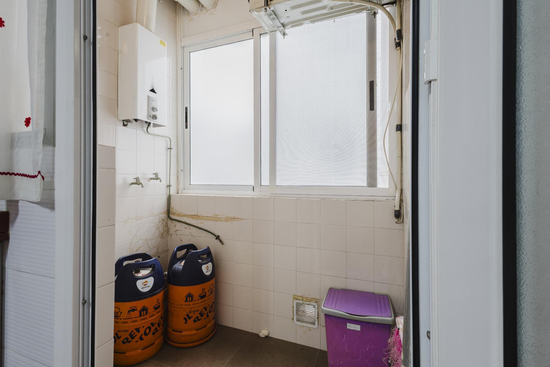 Utility room with two orange gas cylinders on the floor, white tiled walls, a wall boiler, and a purple storage bin near the window.